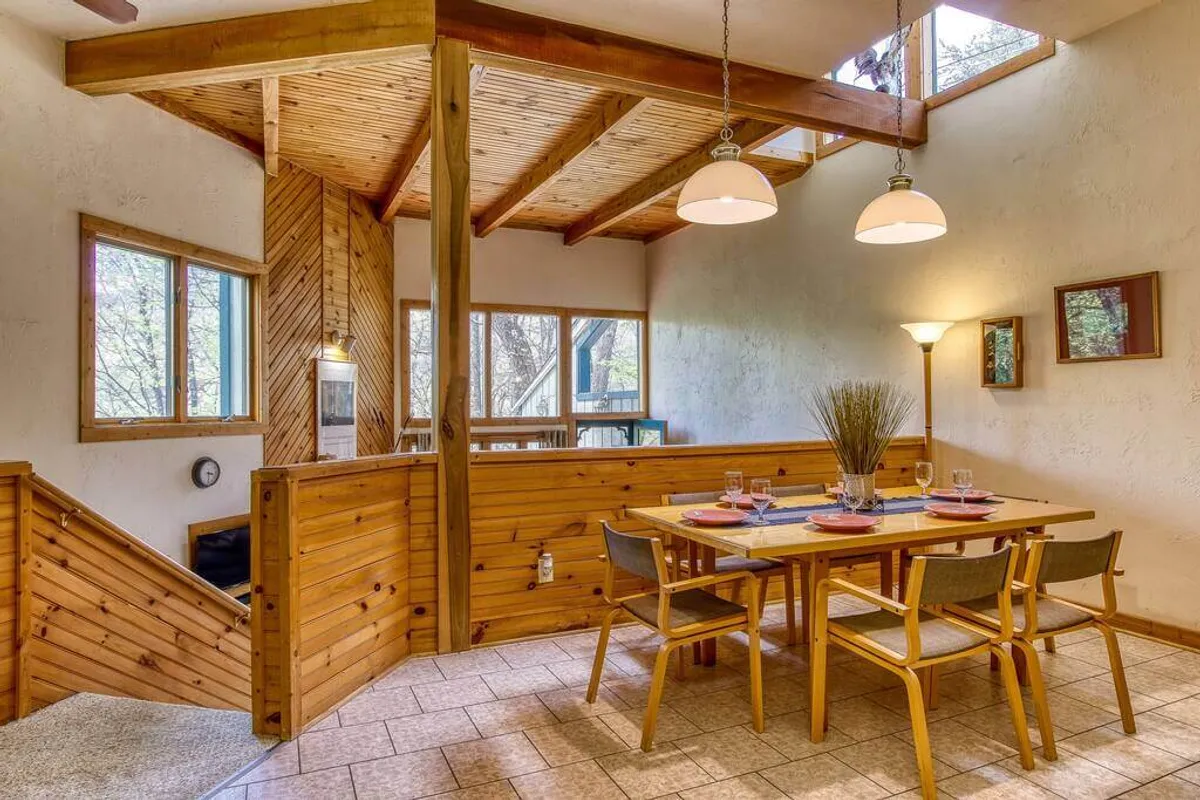 A spacious dining area with a sturdy wooden table, exposed beams, large windows letting in natural light, and decorative wall art. The scene is set for family meals, celebrations, and shared moments, with the openness bridging the living and dining areas.