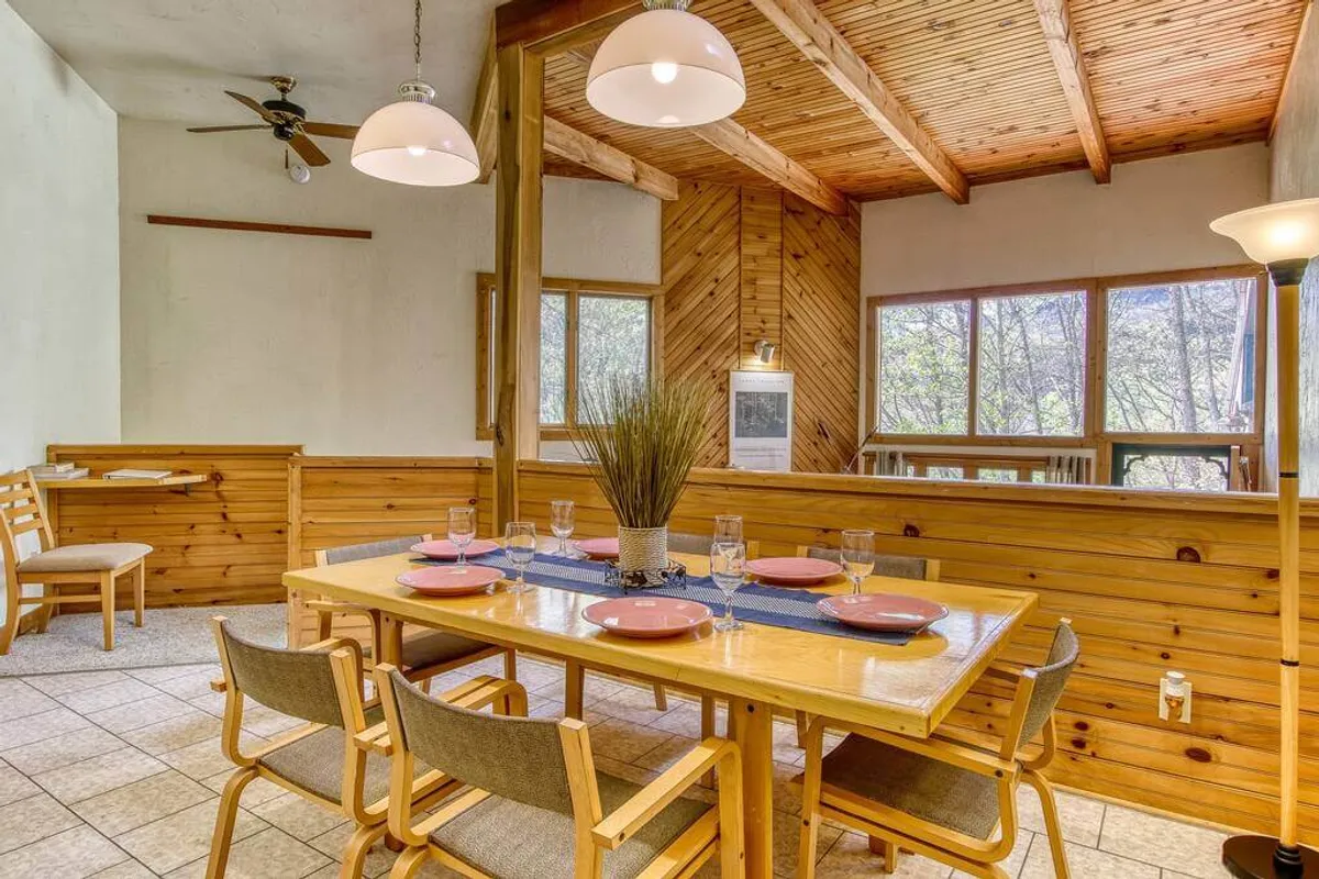 A cozy dining area featuring a spacious table, a wooden ceiling, and large windows, creating an inviting space for casual gatherings or memorable holiday dinners. The setting is intimate and filled with natural light.