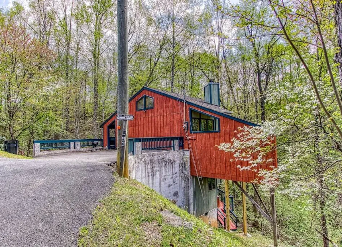 The hero image showcases the cabin's striking, vibrant red exterior and unique architectural design, nestled amidst lush greenery. The setting radiates peace and seclusion, with a welcoming porch perfect for enjoying sunsets and the fresh mountain air. The surrounding woods suggest opportunities for hiking and connecting with nature, while the cabin itself stands as an inviting retreat for families or couples.