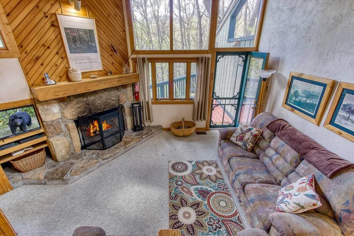 This image showcases a welcoming living room with a stone fireplace, a plush couch, a decorative rug, and large windows that allow natural light to pour in. The space radiates warmth and comfort, ideal for gathering, storytelling, and relaxing. The visible windows also frame the scenic outdoor views, blurring the lines between inside and out.