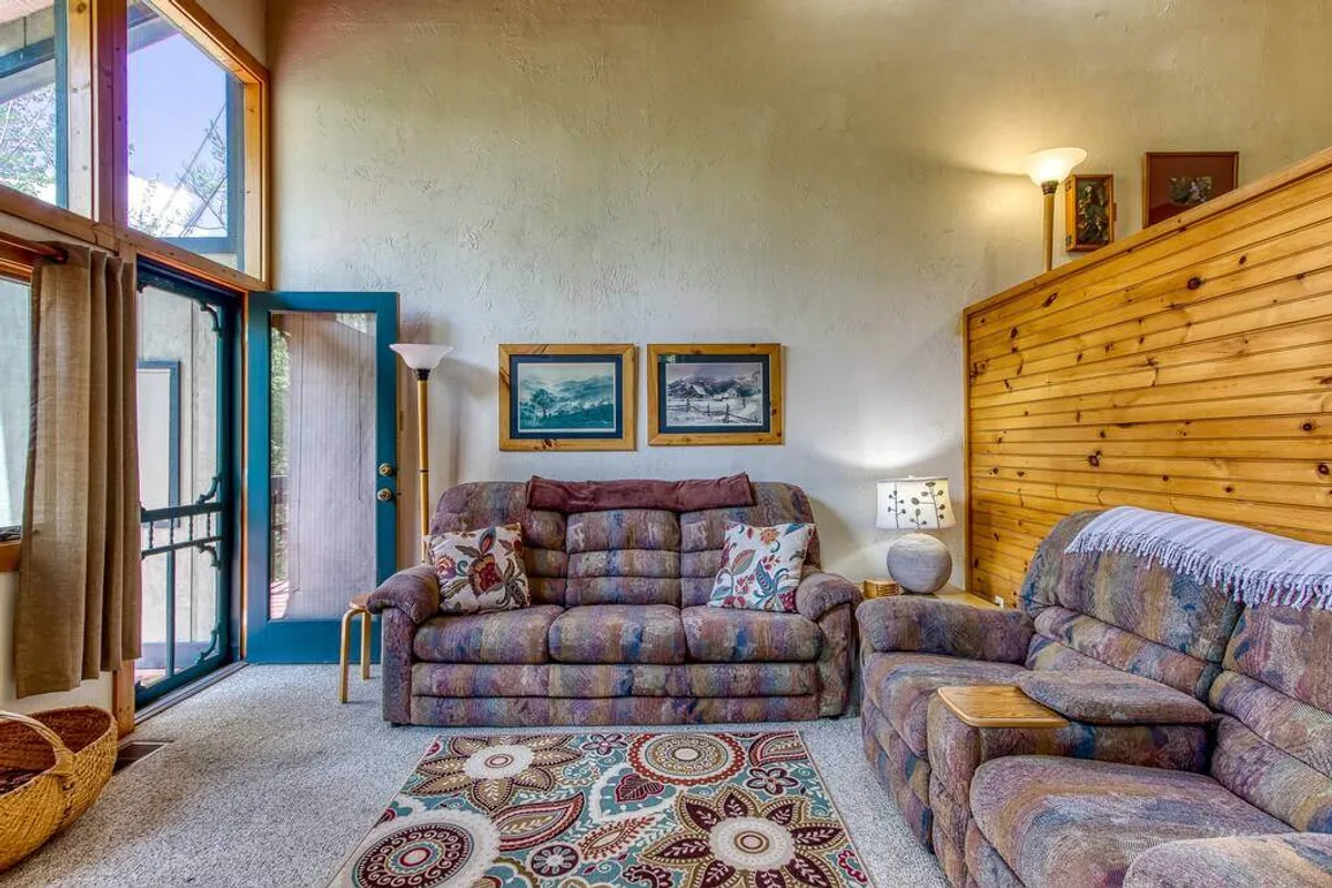 A cozy living space featuring multiple sofas, a colorful rug, wood-paneled walls, framed pictures, and a soft lamp. The arrangement encourages intimate conversations, game nights, or quiet reading sessions, embodying a relaxed and homey feel.