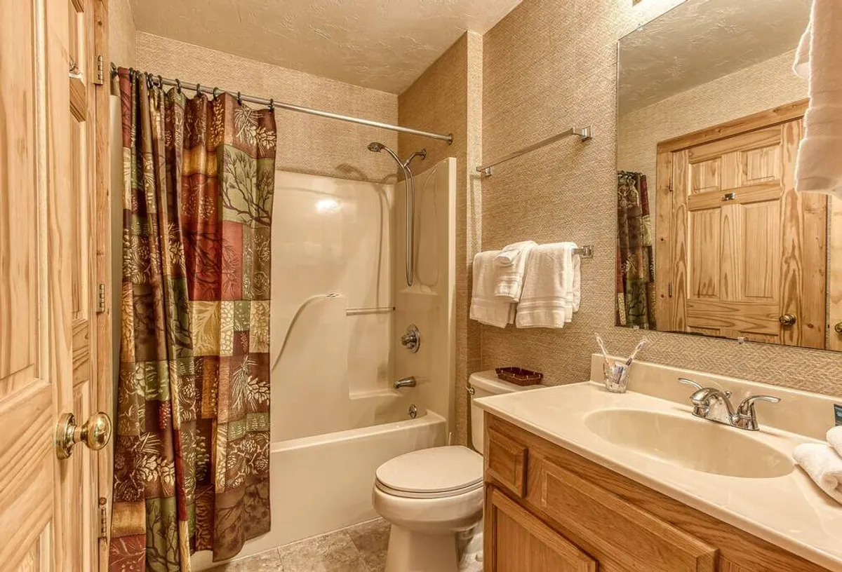 A well-appointed bathroom featuring a soothing bathtub, shower curtain, sink with wooden cabinetry, and neatly arranged towels. The space feels comfortable and practical, providing the ideal setting for unwinding after mountain adventures.