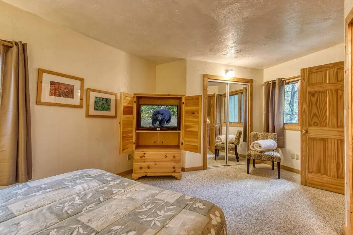 A second inviting bedroom with floral bedding, a wooden TV stand, seating area with a chair, framed artwork, and a large window letting in abundant natural light. The mood is warm and tranquil, perfect for relaxation or quiet downtime.