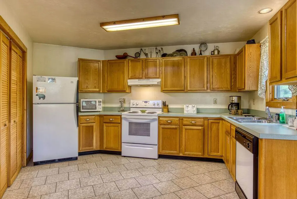 A warm, homey kitchen featuring wooden cabinets, white appliances, a spacious kitchen counter, a sink, and a window with a curtain that allows natural light to fill the space. The layout invites communal meal preparation and casual conversation.