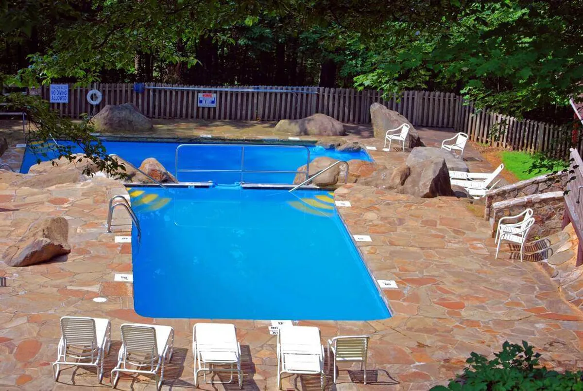 A vibrant community pool area framed by natural rocks and dotted with lounge chairs, where people are swimming, sunbathing, and socializing. The scene is lively and inviting, with a cheerful, resort-like ambiance, highlighting a space perfect for family fun and relaxation.